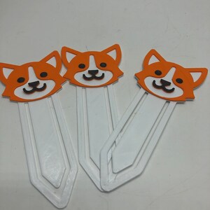 May include: Three orange and white dog-shaped bookmarks with black eyes and noses. The bookmarks are made of a white plastic material and have a rounded shape.