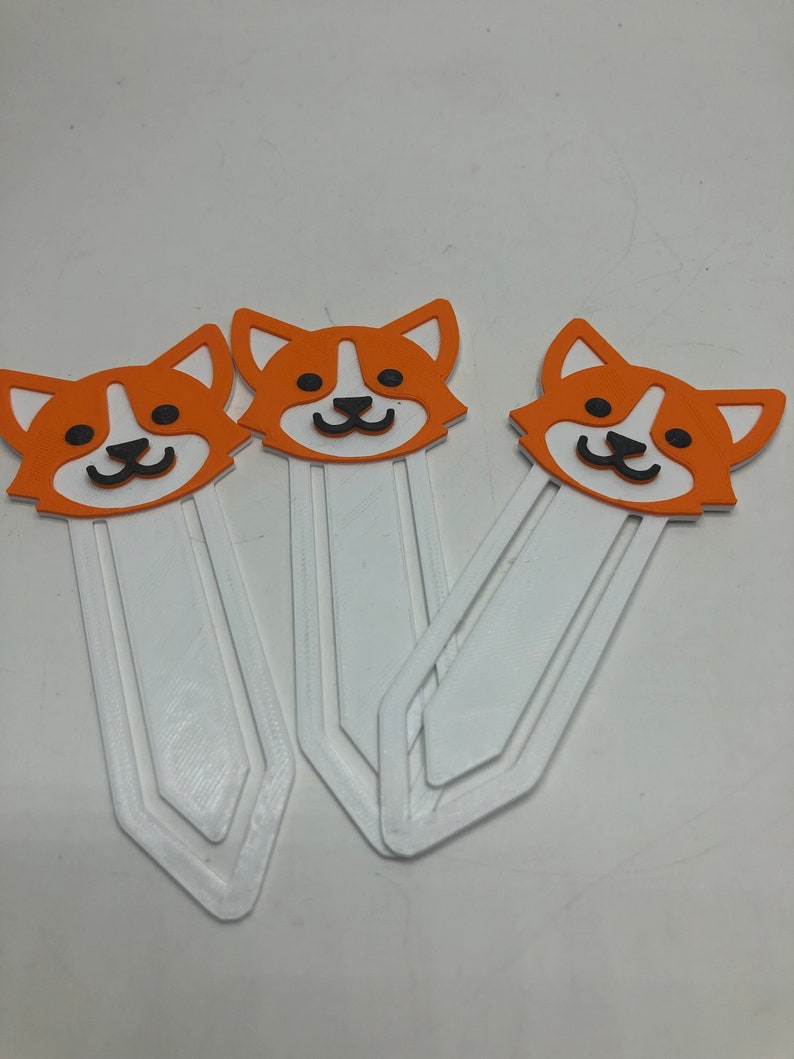 May include: Three orange and white dog-shaped bookmarks with black eyes and noses. The bookmarks are made of a white plastic material and have a rounded shape.