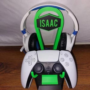 May include: A green and black gaming headset stand with the name "ISAAC" printed on it. The stand is holding a white wireless gaming headset and a white PlayStation 5 controller.