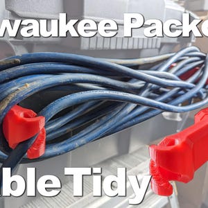 May include: A red plastic cable organiser mounted on a Milwaukee Packout system, holding a coiled blue electrical cord. The organiser features two distinct red brackets attached to the vertical metal rails, with the text Milwaukee Packout Cable Tidy.
