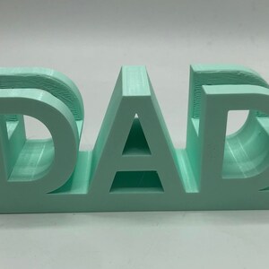 May include: A light mint green 3D printed sign featuring the word DAD in bold, block capital letters. The object has a flat base and a smooth, matte finish, with visible horizontal layer lines characteristic of the additive manufacturing process.