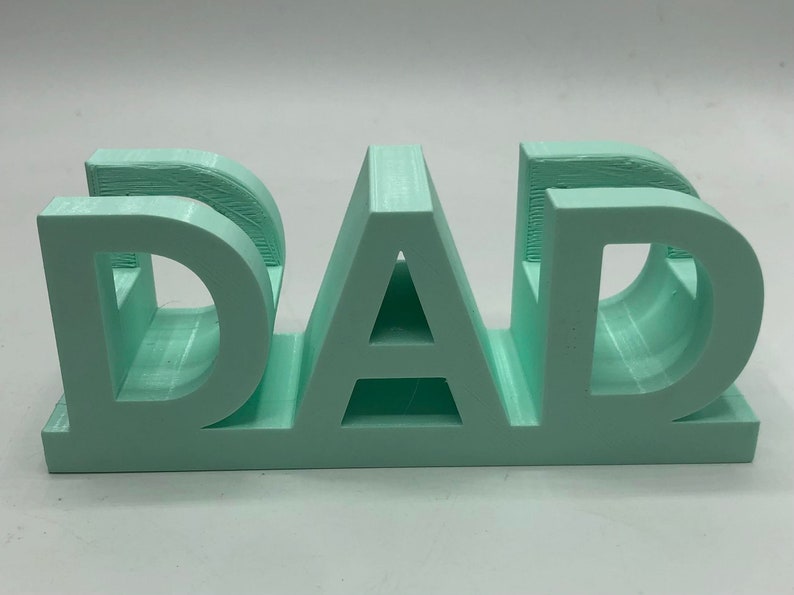 May include: A light mint green 3D printed sign featuring the word DAD in bold, block capital letters. The object has a flat base and a smooth, matte finish, with visible horizontal layer lines characteristic of the additive manufacturing process.