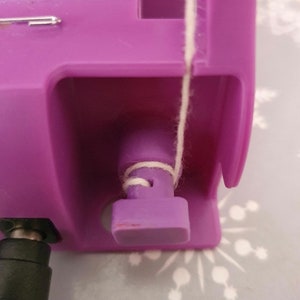 May include: A close-up of a purple plastic yarn dispenser with a white string wrapped around the spool. The dispenser has a small, rectangular opening on the side.
