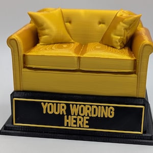 May include: A miniature gold-colored couch trophy with two pillows. The couch sits atop a black base with a gold-framed plaque that reads "YOUR WORDING HERE". A decorative item.