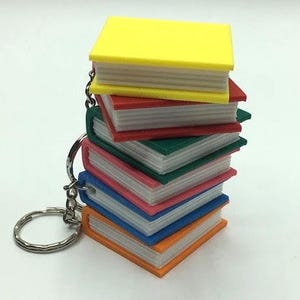 May include: A stack of colourful, miniature book-shaped keyrings with a silver key ring. The books are stacked in order of size, with the largest book on top and the smallest book on the bottom. The books are yellow, red, green, blue, pink, and orange.