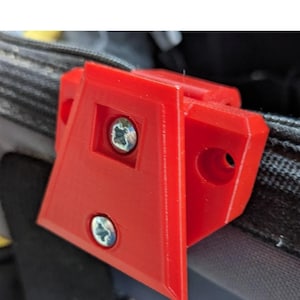 May include: A vibrant red, 3D-printed corner bracket, secured with two silver screws. The bracket is attached to a black and grey textured surface, possibly a bag or case. The design is a right-angle shape with a hole.