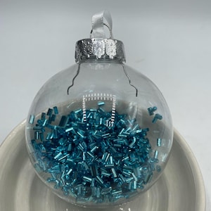 May include: A clear, spherical glass ornament filled with small, metallic turquoise blue cylindrical beads. The ornament features a silver-colored metal cap with a white ribbon loop attached to the top for hanging purposes.