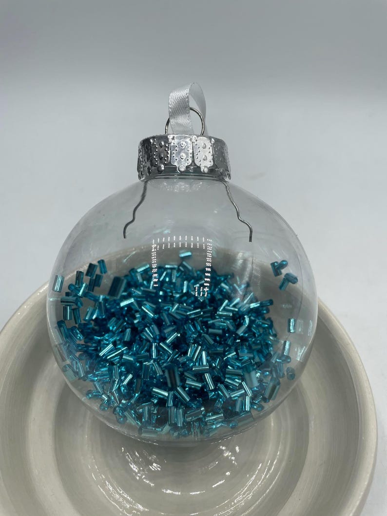 May include: A clear, spherical glass ornament filled with small, metallic turquoise blue cylindrical beads. The ornament features a silver-colored metal cap with a white ribbon loop attached to the top for hanging purposes.