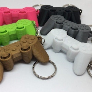 Peut inclure: Cinq porte-clés de manette de jeu en plastique imprimés en 3D de différentes couleurs. Les porte-clés sont en rose, vert, marron, gris et blanc.