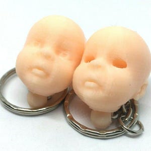 May include: Two small, light pink, 3D printed baby heads attached to silver keychains. The heads are facing forward and have closed eyes.