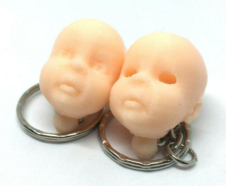 May include: Two small, light pink, 3D printed baby heads attached to silver keychains. The heads are facing forward and have closed eyes.