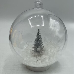May include: A clear, spherical, hollow plastic ornament featuring a small, silver-toned, glittery evergreen tree standing in a base of white, textured, snow-like material. The ornament has a loop at the top for hanging and a flat base.