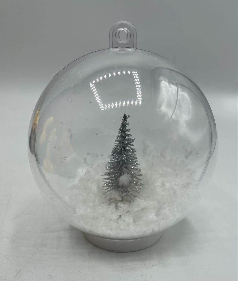 May include: A clear, spherical, hollow plastic ornament featuring a small, silver-toned, glittery evergreen tree standing in a base of white, textured, snow-like material. The ornament has a loop at the top for hanging and a flat base.