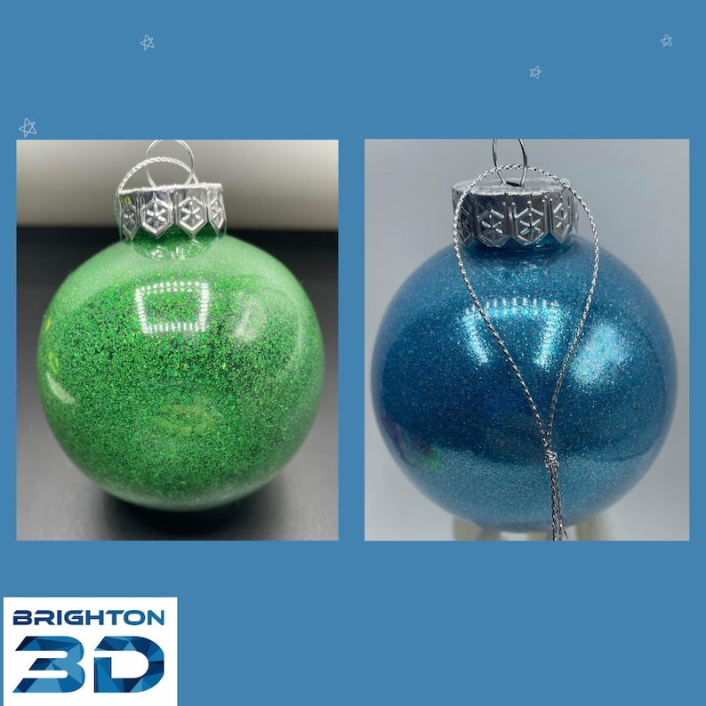 May include: Two spherical ornaments, one green and one blue, both featuring a glittery finish and a silver-toned cap with a snowflake pattern. Each ornament includes a silver hanging string attached to the top for display purposes.