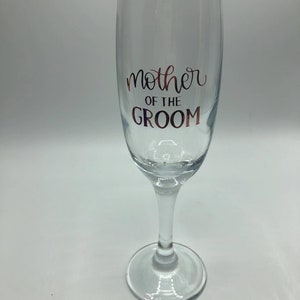 May include: A clear glass champagne flute with the text "mother of the groom" in rose gold lettering.