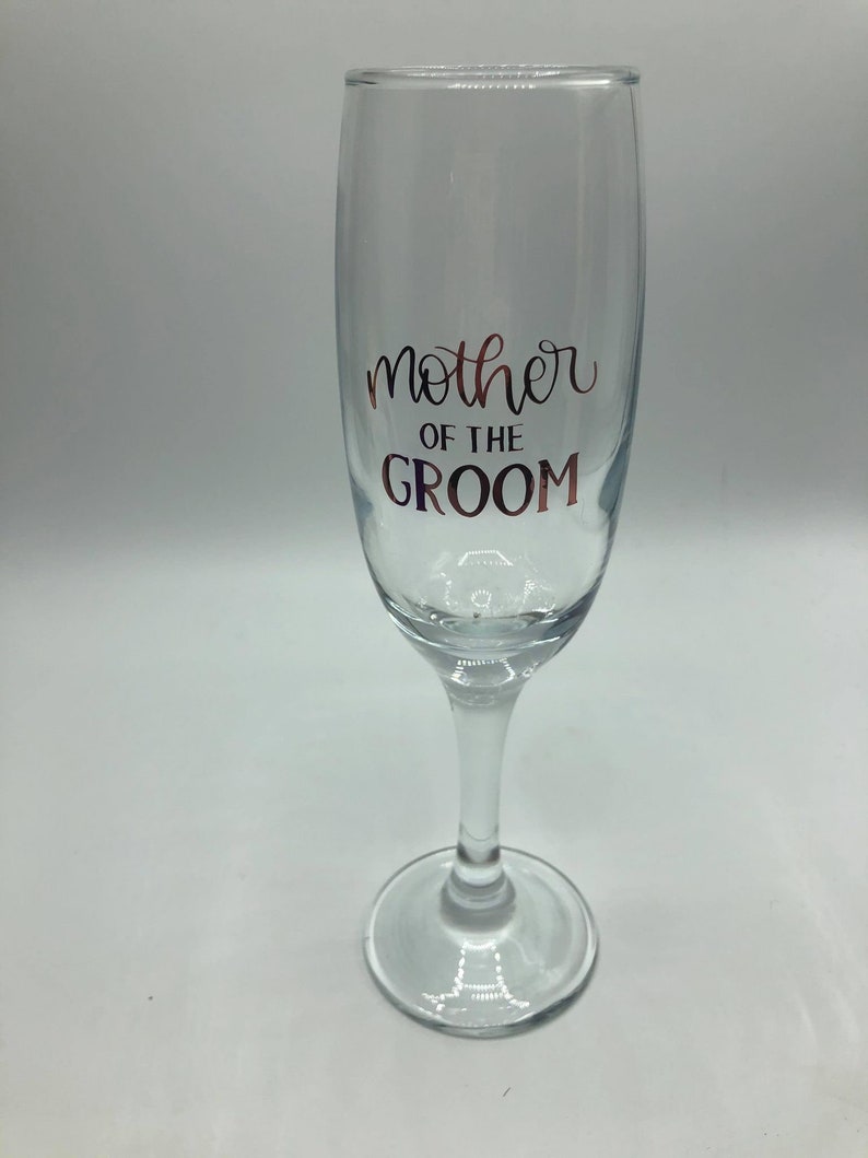 May include: A clear glass champagne flute with the text "mother of the groom" in rose gold lettering.