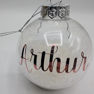 May include: A clear, spherical glass Christmas ornament filled with white, snow-like material. The name Arthur is written in metallic rose gold script on the front. It features a silver metal cap with a snowflake pattern and a silver hanging string.