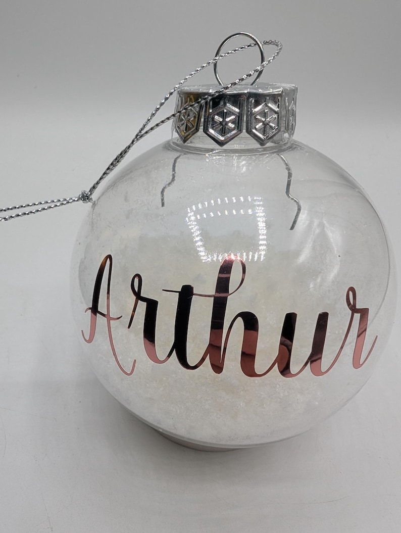 May include: A clear, spherical glass Christmas ornament filled with white, snow-like material. The name Arthur is written in metallic rose gold script on the front. It features a silver metal cap with a snowflake pattern and a silver hanging string.