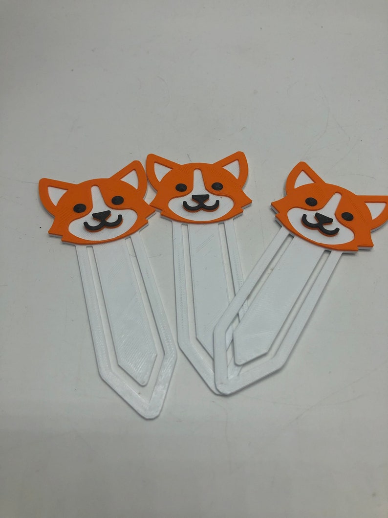 May include: Three white plastic bookmarks with orange and black cartoon dog faces. The dog faces have white eyes and black noses and mouths.