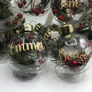 May include: Clear glass spherical ornaments with gold metallic caps, each containing green pine needles, small brown pinecones, and red berries. The ornaments feature personalized names written in gold cursive script, including Emma, Ben, and Tom.