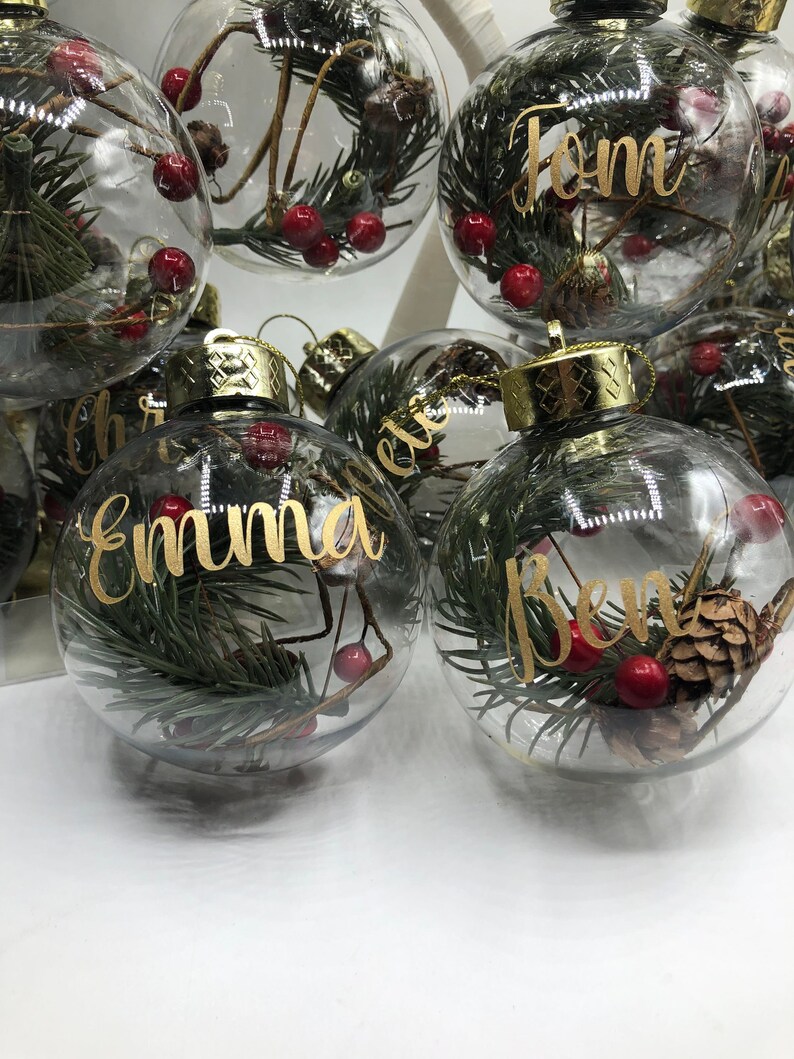 May include: Clear glass spherical ornaments with gold metallic caps, each containing green pine needles, small brown pinecones, and red berries. The ornaments feature personalized names written in gold cursive script, including Emma, Ben, and Tom.
