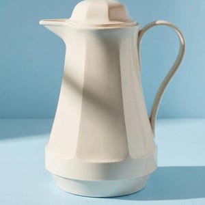 Könnte beinhalten: Eine cremefarbene Thermokaraffe mit facettiertem, geometrischem Körper, passendem Deckel mit sechseckigem Oberteil und einem geschwungenen Griff. Die Kanne hat einen breiten Boden, der sich nach oben zu einem spitzen Ausguss verjüngt.