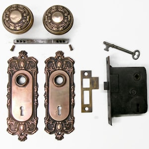 May include: Antique door hardware set featuring two ornate bronze doorknobs, two matching backplates, a lock mechanism, a strike plate, a key, and screws. The hardware has a detailed, vintage design with a copper and bronze finish.