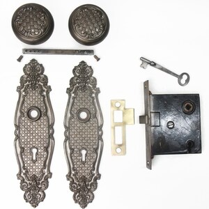 May include: Antique door hardware set, featuring two ornate door knobs with starburst patterns, two decorative backplates, a lock, key, and strike plate. The set is made of metal with a dark, aged finish.
