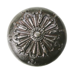 May include: A round, antique-style metal button with an ornate floral design. The button is dark gray with a slightly textured surface and a raised, detailed pattern in a lighter gray color. The central design is a circle with radiating petal-like shapes.