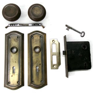 May include: Antique door hardware set featuring two round bronze doorknobs, two rectangular brass plates, a black metal lock, a key, and other components. The set includes all the necessary parts for a complete door installation.
