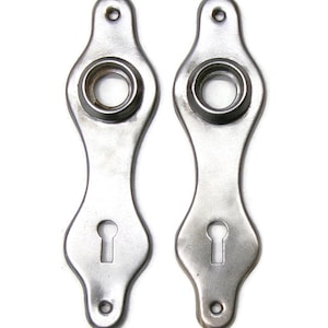May include: Two antique silver-toned door escutcheons. Each escutcheon has a keyhole, a circular opening, and a hole at the top and bottom for mounting. The escutcheons have a decorative, elongated shape.