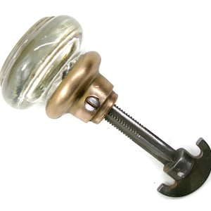 May include: A clear glass door knob with a rounded, smooth surface attached to a metallic, bronze-toned base. A long, threaded metal spindle extends from the base, ending in a dark, semi-circular metal mounting plate with a small screw visible.