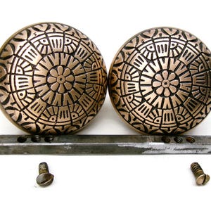 May include: Two antique bronze-colored door knobs with an intricate circular design. The knobs are round with a central flower pattern and geometric shapes. Includes a metal bar and screws for installation.