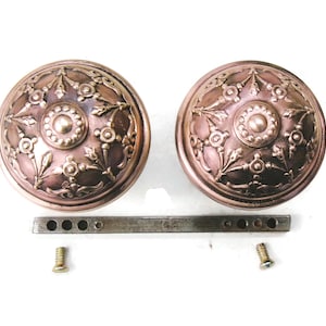 May include: Two antique bronze door knobs with intricate floral and leaf designs. The knobs are round with a central circular detail and are accompanied by a metal bar and screws, suggesting a complete set for installation.