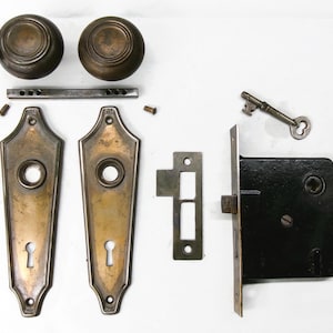 May include: Antique door hardware set, including two bronze doorknobs, two ornate backplates, a mortise lock, a key, and other components. The hardware has a vintage aesthetic.