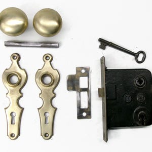 May include: An antique door hardware set featuring two brass doorknobs, a key, a black lock, two brass escutcheons, a strike plate, and a connecting rod. The hardware has a vintage aesthetic.