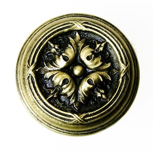 May include: A round, antique brass-colored button with an ornate floral design. The button features a raised, detailed pattern of stylized leaves and swirls within a circular frame. The button is set against a white background.
