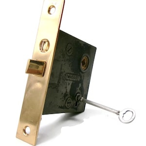 May include: A vintage Corbin door lock with a brass-colored faceplate and a dark metal body. A small key is inserted into the lock. The lock has a rectangular shape with rounded edges and visible screw heads.