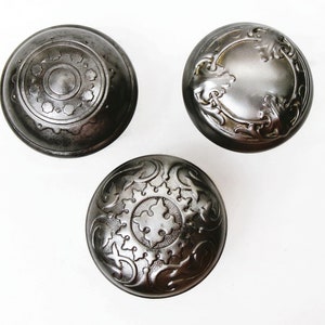 May include: Three antique, dark metal cabinet knobs with intricate embossed designs. One knob features a central circle with floral accents, another has a floral border, and the third displays a starburst pattern. The knobs are set against a white background.
