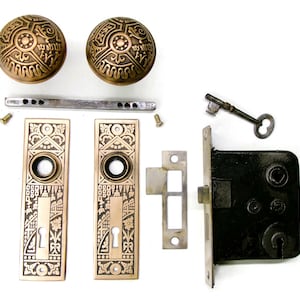 May include: A vintage door hardware set featuring two round brass knobs with intricate embossed patterns, two matching rectangular brass backplates with keyholes, a metal spindle, two small screws, a strike plate, a mortise lock, and a skeleton key.
