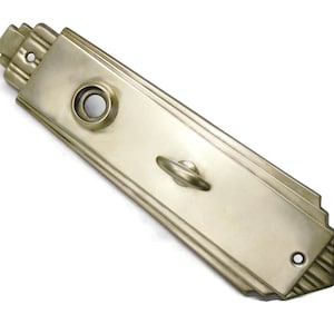 May include: A rectangular, Art Deco-style door plate in a muted gold tone. The plate features a circular keyhole, a small handle, and decorative linear detailing. The design includes stepped edges and a smooth, metallic finish.