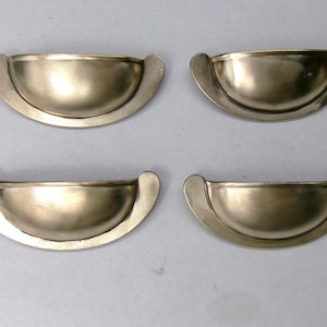 May include: Four antique brass-toned half-moon drawer pulls. Each pull has a curved, bowl-like design with rounded ends. The pulls are arranged in two rows of two against a neutral background. These are likely for furniture restoration.