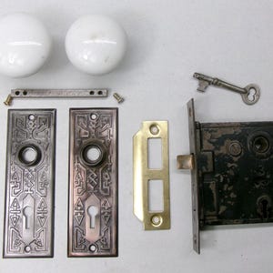 May include: An antique door hardware set, featuring two white porcelain doorknobs, decorative metal plates, a key, and a black lock mechanism. The set includes a brass strike plate and various screws and connecting pieces.