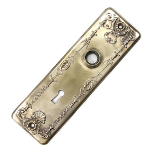May include: Antique brass-colored door plate with ornate floral and scrollwork detailing. The rectangular plate features a keyhole and a circular opening, with decorative borders and corner accents. A vintage-style hardware piece.