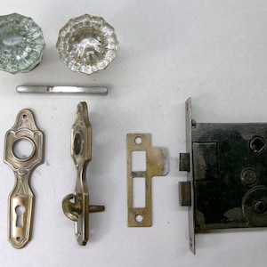 May include: Antique door hardware set featuring two clear glass doorknobs, a threaded rod, and various brass and metal components, including a strike plate and a mortise lock. The set is designed for vintage doors.