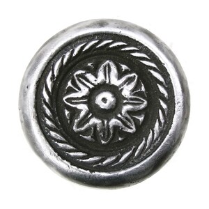 May include: A circular silver-toned button featuring a raised central star-shaped floral motif surrounded by a textured rope-like border. The background of the design is darkened to provide contrast against the polished metallic surface of the button.