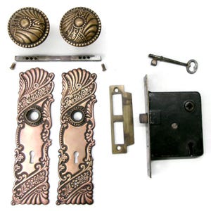 May include: Antique door hardware set featuring two ornate bronze doorknobs, two copper-toned door plates, a key, a latch, and a strike plate. The set includes screws and a connecting bar. The hardware has an aged, vintage appearance.