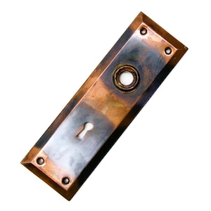 May include: Rectangular antique door plate with a keyhole and a circular opening. The plate is made of copper and has a tarnished, aged appearance. Four screw holes are visible in the corners. The door plate is a vintage hardware item.