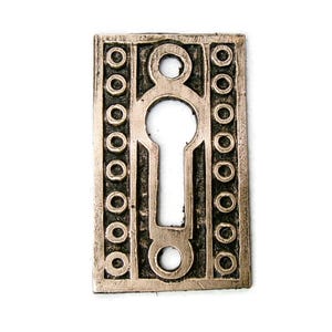 May include: A rectangular, antique-style keyhole cover in a bronze finish. The cover features a keyhole shape in the center, surrounded by a decorative border of small circles. The overall design is symmetrical and detailed, suggesting a vintage or historical aesthetic.