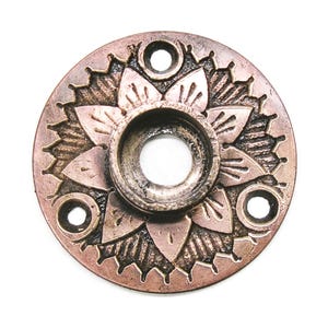 May include: A copper-colored, circular decorative plate with a floral design. The plate has a central hole and three smaller holes evenly spaced around the edge. The floral pattern is embossed, adding texture to the metal.